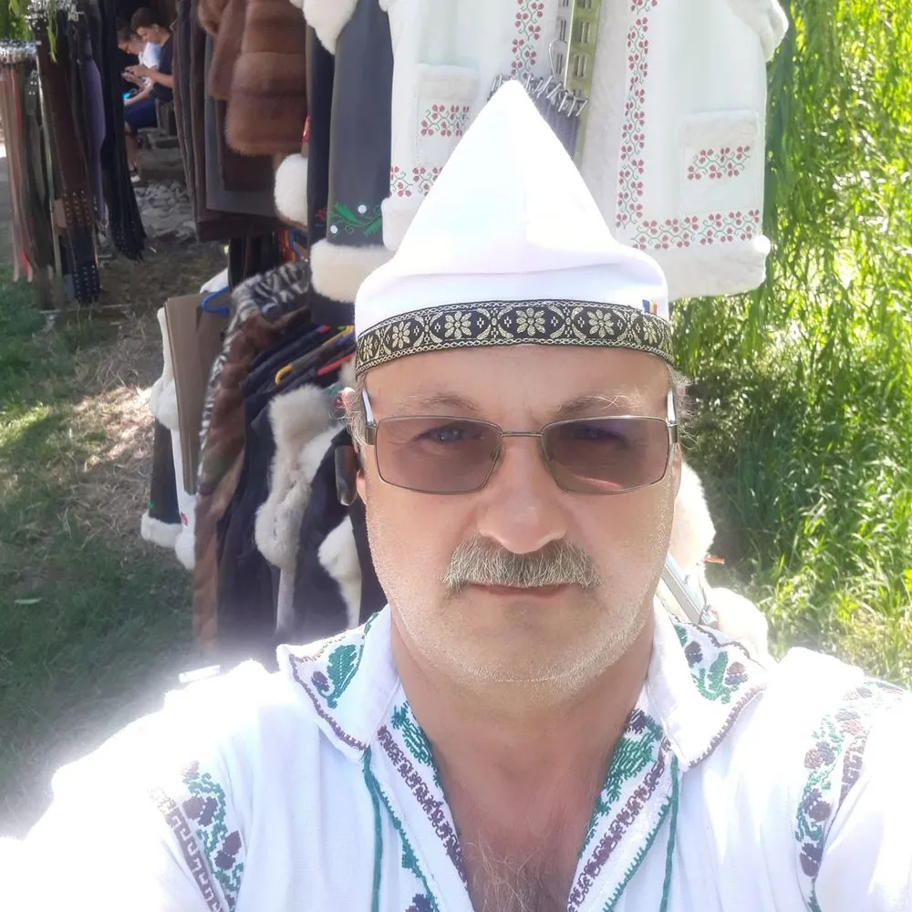 Meșterul la un târg în aer liber, cu o căciulă înaltă și țuguiată pe cap și cămașă populară brodată, lângă standul cu curele și cojoace.