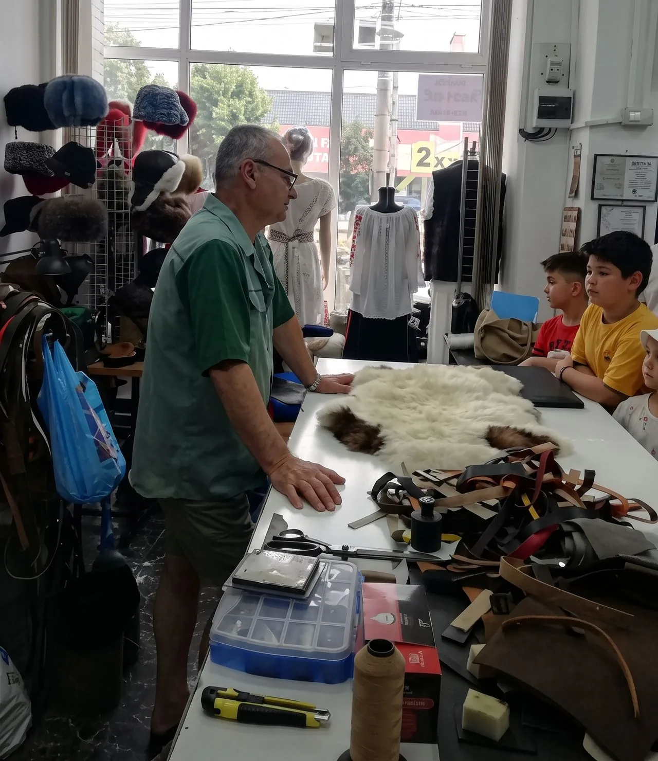 Mâinile meșterului așezate lângă o piele de miel întinsă pe masa de croit, în timpul explicației.